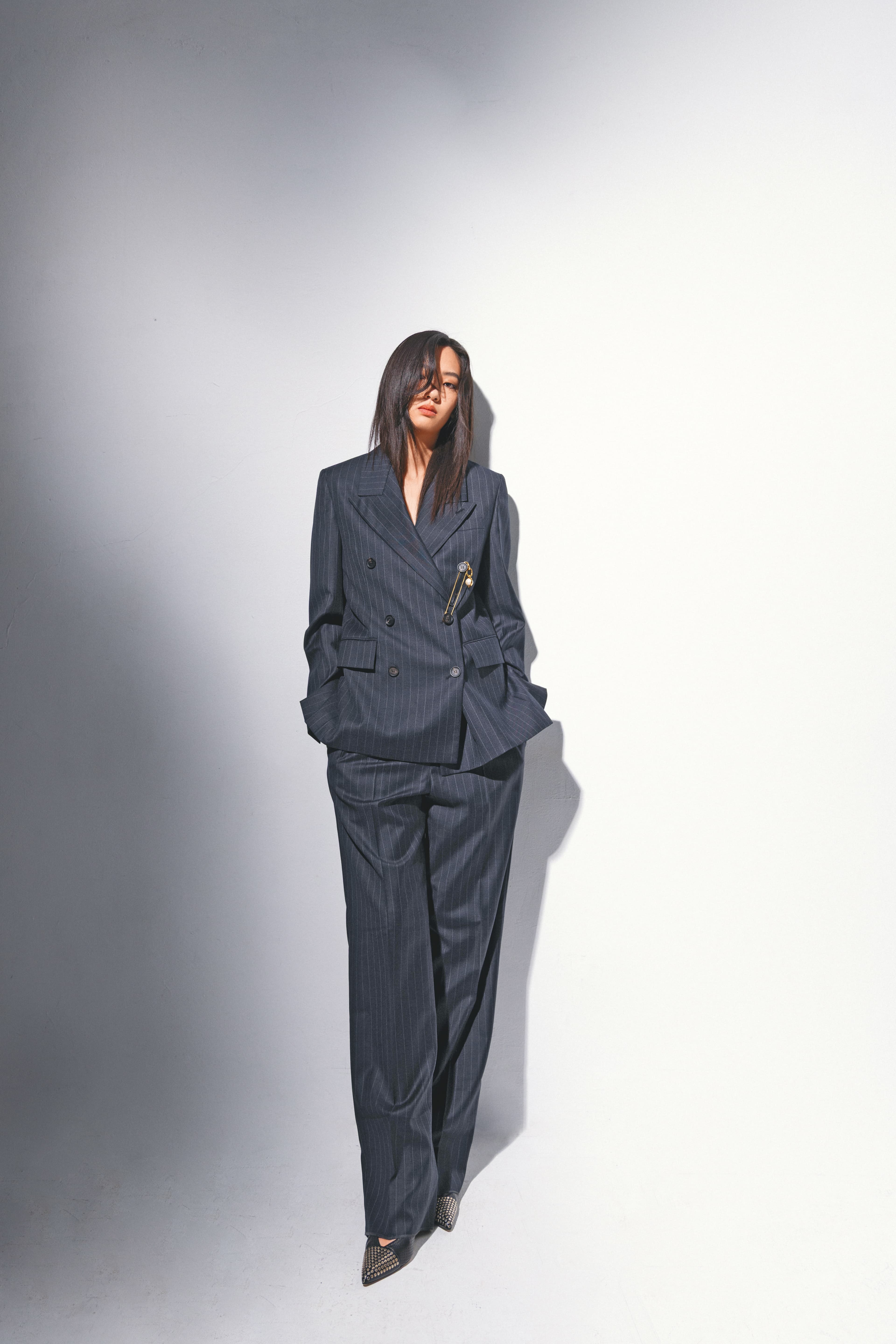 炭灰色細條紋羊毛西裝外套 NT$89,900 by FERRAGAMO；炭灰色細條紋羊毛西裝褲 NT$41,900 by FERRAGAMO；尖頭高跟鞋 價格店洽 by Chloé；CELINE CHARMS金色飾面安全扣針胸針 NT$11,500 by CELINE；CELINE CHARMS PEARL吊飾 NT$11,500 by CELINE。