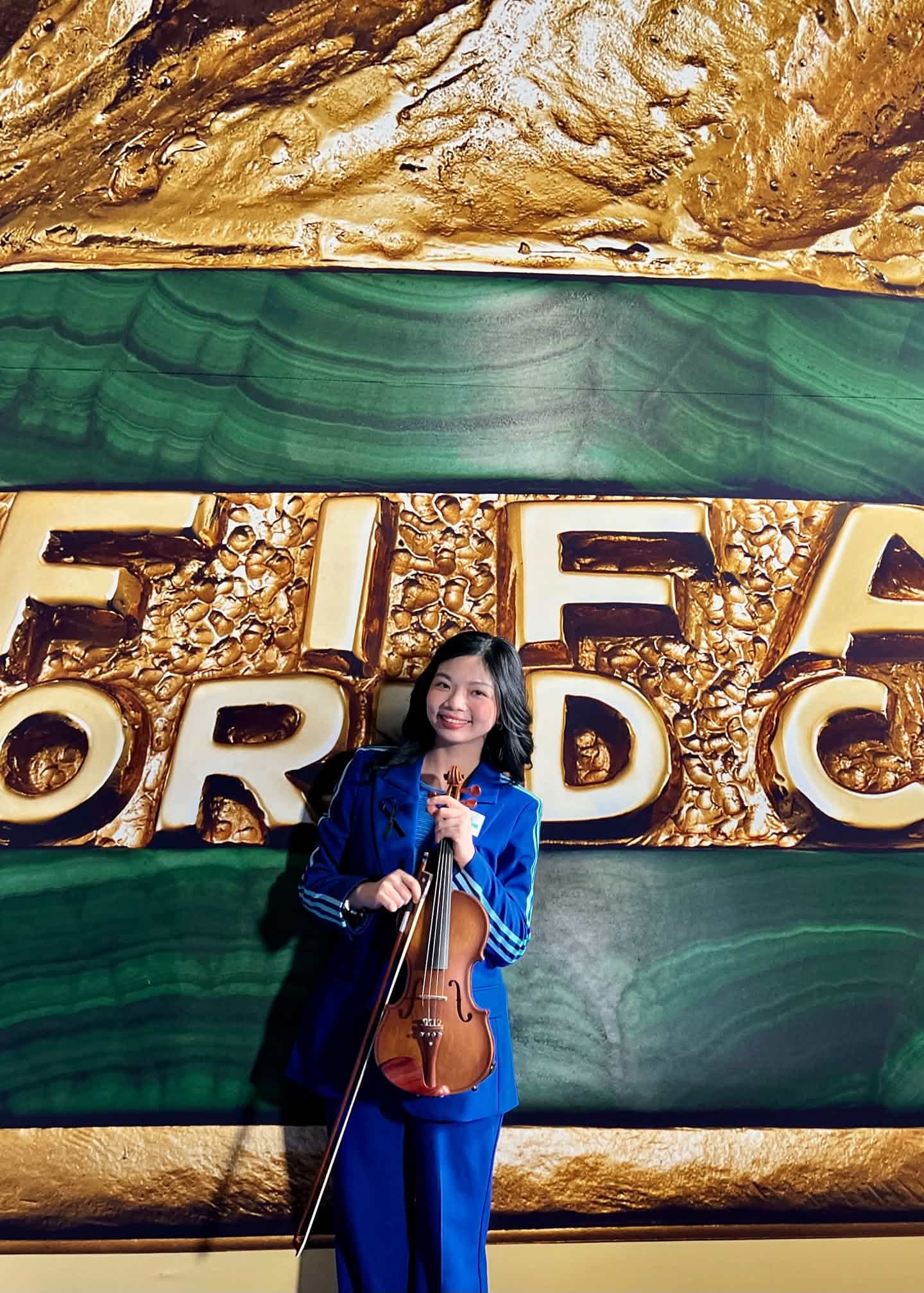 黃薏蓉拿著小提琴,在FIFA World Cup背板前留下紀念照片。(黃薏蓉提供)